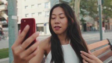 Şehir parkında bankta otururken cep telefonundan selfie çeken genç bir kadın.