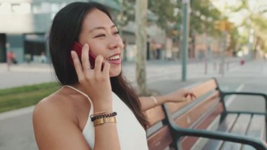 Genç bir kadın bankta oturuyor ve cep telefonuyla konuşuyor.