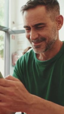 Orta yaşlı bir adam kafede oturup cep telefonundan video izliyor.