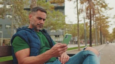 Orta yaşlı erkek turist park bankında otururken cep telefonu kullanıyor