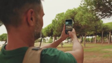 Yakın plan, orta yaşlı adam şehir parkında dururken akıllı telefondan fotoğraf çekiyor.