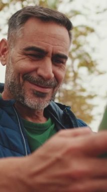 Yakın plan, orta yaşlı bir adam parkta bankta oturuyor ve cep telefonu kullanıyor.