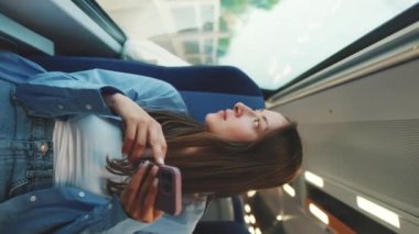 Dikey video, uzun kahverengi saçlı, mavi gömlekli tatlı kız, banliyö treniyle seyahat ederken pencereden dışarı bakıyor, akıllı telefon ekranında kaydırıyor.