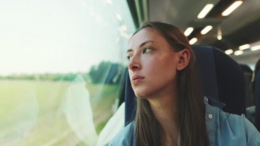 Uzun kahverengi saçlı, mavi gömlekli hoş bir kız tren yolculuğu sırasında pencereden dışarı dikkatlice bakıyor.