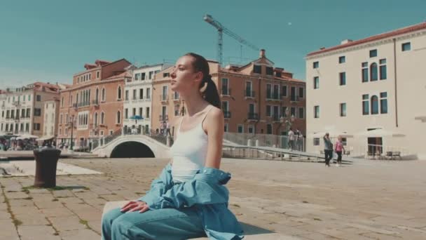 長い茶色の髪をした美しい女の子 青いシャツを着て ヴェネツィアの堤防上に座っています — ストック動画