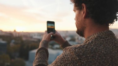 Mutlu adam şafakta balkonda duruyor, fotoğraf çekiyor, akıllı telefondaki güzel manzaranın videosunu çekiyor.