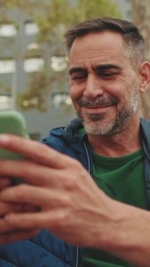 Yakın plan, orta yaşlı bir adam parkta bankta oturuyor ve cep telefonu kullanıyor.