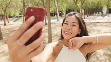 Beyaz bluz ve kot giyen gülümseyen esmer kız, şehir parkında dururken cep telefonundan selfie çekiyor.