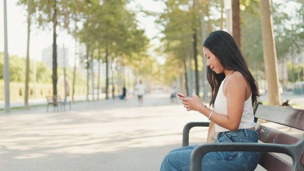Симпатичная брюнетка, в белом топе и джинсах, сидит на скамейке в парке со смартфоном в руках, печатает сообщение