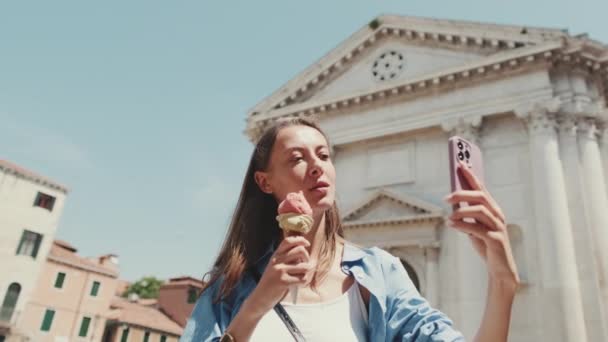美丽的女孩 棕色长发 身穿蓝色衬衫 站在老城区广场时 喜欢吃冰淇淋 还喜欢用手机自拍 — 图库视频影像