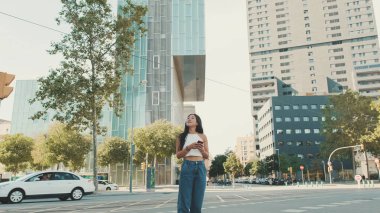 Beyaz bluz ve kot giyen sevimli mutlu kız modern şehir arka planında dururken cep telefonunda harita uygulaması kullanıyor.