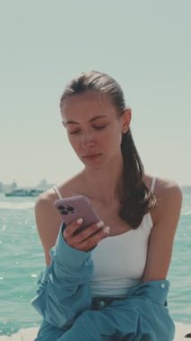 Dikey video, uzun kahverengi saçlı, mavi tişörtlü güzel kız, Venedik 'te deniz kenarında oturuyor ve arka planda cep telefonu, tekne ve sürat teknesi kullanıyor.