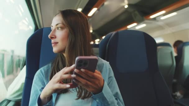 青いシャツを着た長い茶色の髪のかわいい女の子は 郊外の電車で旅行中に窓の外を見て スマートフォンの画面上でスクロールします — ストック動画