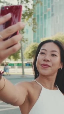 Gülümseyen genç bir kadın modern şehir arka planında dururken cep telefonuyla selfie çekiyor.