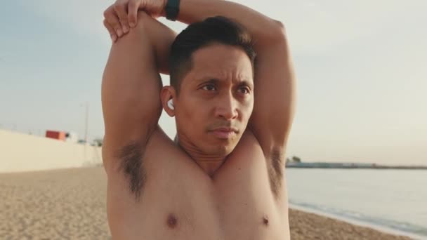 トレーニングの前に腕の筋肉を伸ばしてビーチに立つ若い運動選手が — ストック動画