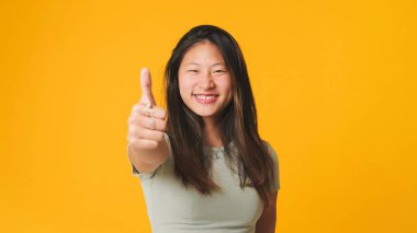 Gri elbiseli kız kameraya bakıyor, gülüyor ve sarı arka planda izole edilmiş parmaklarını gösteriyor.