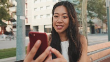 Beyaz bluzlu güzel esmer kız gülümsüyor ve kentin arka planında bankta otururken selfie çekiyor.