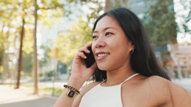 Beyaz bluz ve kot giyen tatlı esmer kız, kentin arka planında bankta oturuyor, akıllı telefondan konuşuyor.