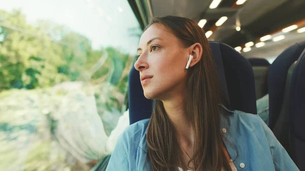 stock image Close up, cute girl with long brown hair, wearing blue shirt, wearing wireless earphone into ear traveling by suburban train looking out the window