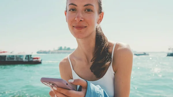 Beautiful girl with long brown hair, dressed in blue shirt, sits on embankment in Venice and uses mobile phone and look at camera, boats and speedboats on background