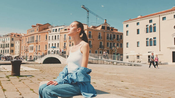 Beautiful girl with long brown hair, dressed in blue shirt, sits on embankment in Venice