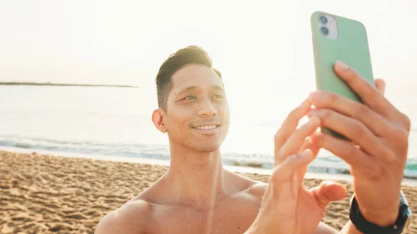 ビーチに立って裸の胴を持つ選手は スマートフォンで海の美しい写真を撮ります — ストック写真