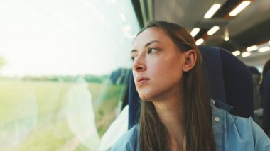 Uzun kahverengi saçlı, mavi gömlekli hoş bir kız tren yolculuğu sırasında pencereden dışarı dikkatlice bakıyor.