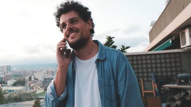 Terasta, arka planda, cep telefonuyla konuşan genç adamın yakın çekimi.