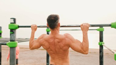 Orta yaşlı kaslı bir adam deniz arka planındaki yatay çubukta egzersiz yapıyor.