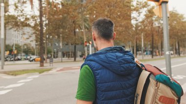 Sırt çantalı, günlük giysiler giyen yetişkin bir turist yaya geçidinde dikilirken etrafına bakıyor.