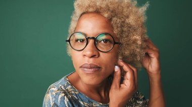 Gözlüklü, mavi tişörtlü, aynadaki gibi kameraya bakan, saçını düzelten, yeşil-mavi arka planda izole edilmiş bir kızın yakın çekimi.