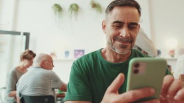 Orta yaşlı bir adam kafede oturup cep telefonundan video izliyor.