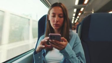 Uzun kahverengi saçlı, mavi gömlekli, banliyö treniyle seyahat eden tatlı bir kız, akıllı telefon ekranında kaydırıyor.