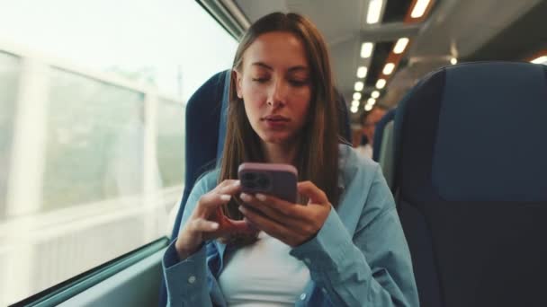 長い茶色の髪をしたかわいい笑顔の女の子 青いシャツを着て 郊外の電車で旅行中 スマートフォンの画面上でスクロールします — ストック動画