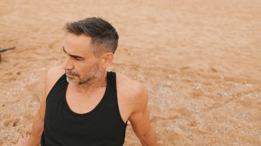 Orta yaşlı atletik adam sahilde otururken antrenmandan sonra dinleniyor.