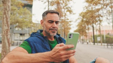 Normal kıyafetler giyen, parkta bankta oturan, akıllı telefondan görüntülü konuşan olgun bir adam.