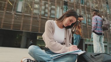 Öğrenci dışarıda otururken ders çalışıyor. Kız sınava hazırlanıyor, not defterine not alıyor.