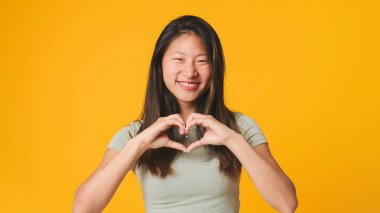 Gri bluzlu kız kameraya bakıyor, gülümseyerek elleriyle kalbi şekillendiriyor. Stüdyoda sarı arka planda izole edilmiş aşk bakımını ifade ediyor.