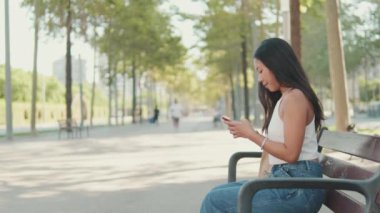 Young woman sitting on city park bench uses mobile phone