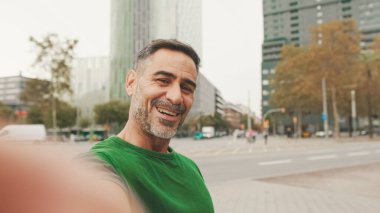 Neşeli, olgun bir adam gündelik kıyafetler giyip modern binalarda cep telefonuyla konuşuyor.