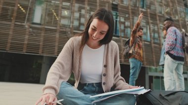 Öğrenci dışarıda otururken ders çalışıyor. Kız sınava hazırlanıyor, not defterine not alıyor.