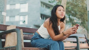 Beyaz bluz ve kot giyen tatlı esmer kız elinde akıllı telefonuyla sokakta bankta oturuyor ve etrafına bakıyor.