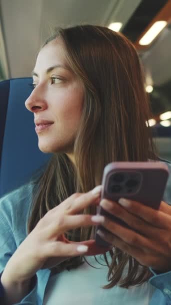 縦型ビデオ 長い茶色の髪のかわいい女の子 青いシャツを着て 郊外の電車で旅行中に窓の外を見る スマートフォンの画面上のスクロール — ストック動画