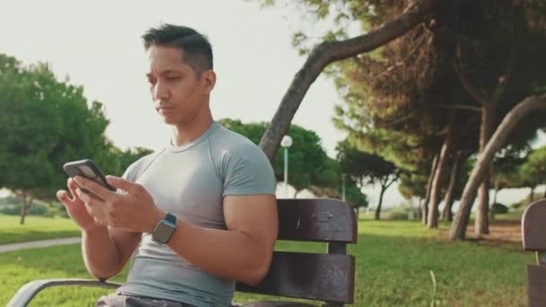 Νεαρός Που Κάθεται Ένα Παγκάκι Του Πάρκου Χρησιμοποιεί Ένα Κινητό — Αρχείο Βίντεο