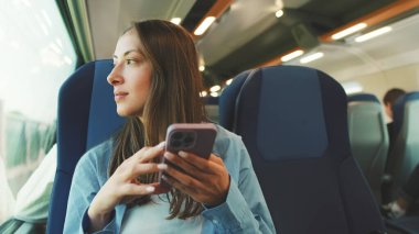 Uzun kahverengi saçlı, mavi gömlekli, banliyö treniyle seyahat ederken pencereden dışarı bakan, akıllı telefon ekranında kaydıran tatlı bir kız.