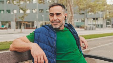 Kablosuz kulaklıkla günlük giysiler giyen olgun bir adam. Parktaki bankta otururken müziğin keyfini çıkartıyor. Alt manzara, yumuşak odaklanma.