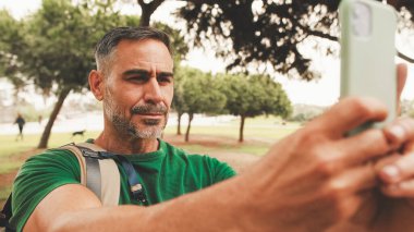 Omzunda sırt çantası olan orta yaşlı bir adam şehir parkında dikilirken cep telefonuyla fotoğraf çekiyor.