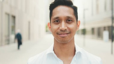 Genç işadamı iş bölgesinde dururken gülümseyip kameraya bakıyor.