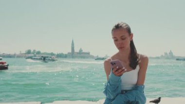 Uzun kahverengi saçlı, mavi gömlekli güzel bir kız, Venedik 'te deniz kenarında oturur ve arka planda cep telefonu, tekne ve sürat teknesi kullanır.