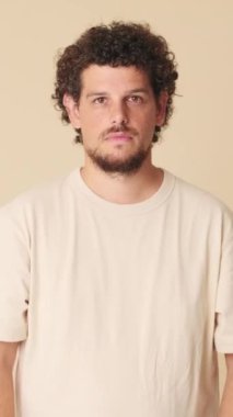 Kollarını kavuşturup kameraya bakan genç bir adam. Stüdyoda bej arka planda izole edilmiş bir gülümsemeyle.